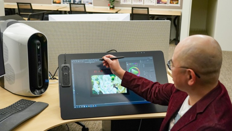In a busy office space, a bald man draws on a computer tablet using a specialized stylus.