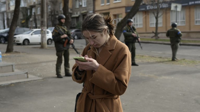 A woman looks at her cellphone near soldiers standing guard outside a government building hit by Russian rockets in Ukraine.