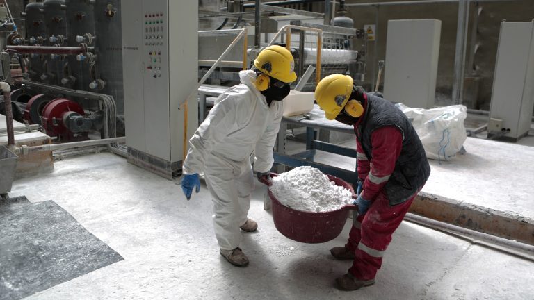 Two people carrying a bucket of white lithium carbonate inside a factory.
