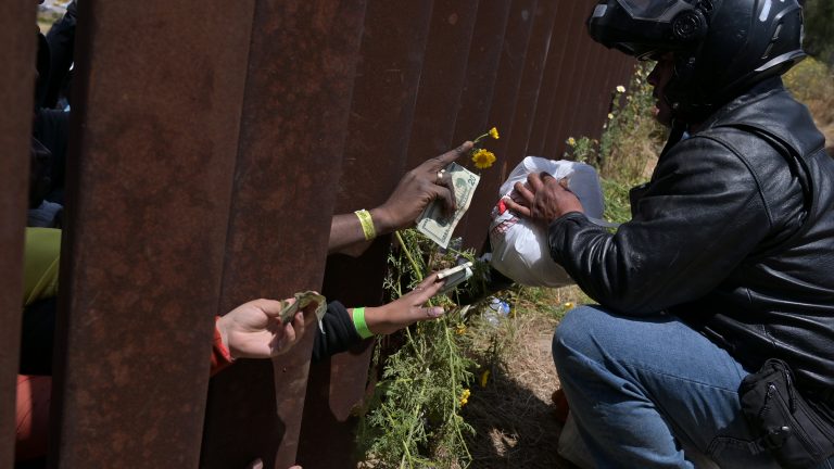 A photo of migrations handing money to a app-based food delivery driver through the United States border wall in Tijuana.