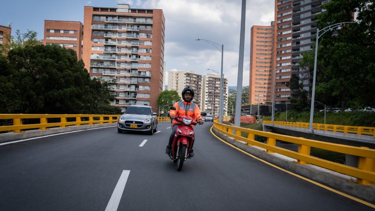 A photo of a delivery rider, wearing an orange jacket, driving a motorbike on a highway in Colombia.