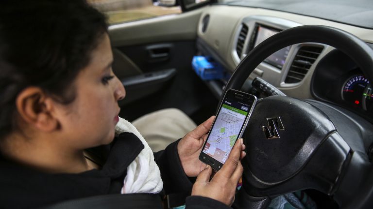A photo showing a woman looking at an app on her phone while seated in the driver's seat of a car.