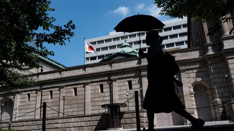A photo showing a silhouette of a person carrying an umbrella past the exterior of a tan, stone building with a Japanese flag flying over it.