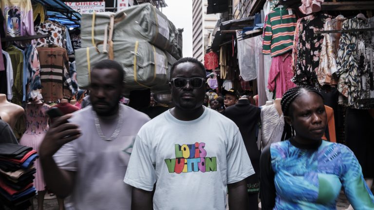 A portrait of a man, standing in the middle of a busy market in Lagos, as other people rush by him in a blur.