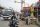 A photo showing a street scene in India with a man riding a motorbike, a golden idol draped in flowers with his hand raised, a bus, and buildings with telephone lines running between them.