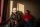 Mauricio Bustos (right) together with his brother Marcelo Bustos (left) who wears a white robotic mask. Both are sitting on the bed in the small room they share in Santiago, Chile, sunlight shining through one side.