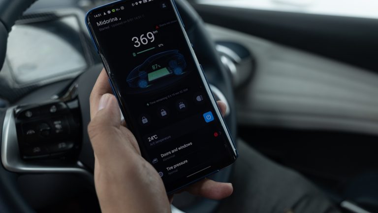 Una fotografia di una mano che tiene un telefono sul sedile del conducente di un veicolo. Lo schermo del telefono mostra la velocità, la temperatura e altri dettagli di un'applicazione per smartphone per un'auto elettrica.