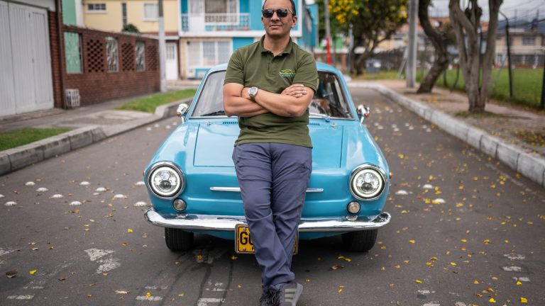 A photograph of a man in a khaki green polo shirt and sunglasses with his arms folded leans against a blue Fiat car parked in the middle of a lane.