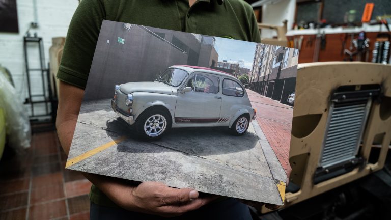 A photograph of a person wearing a polo khaki tshirt holding a printed photo showing a a cream car parked on the street.