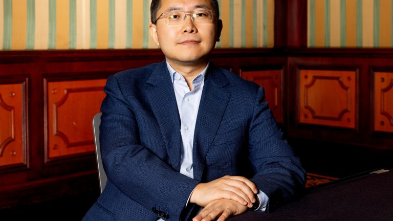 A photograph of President of Alibaba.com, Kuo Zhang, wearing a blue suit and blue dress shirt, seating at a table in an ornate room.