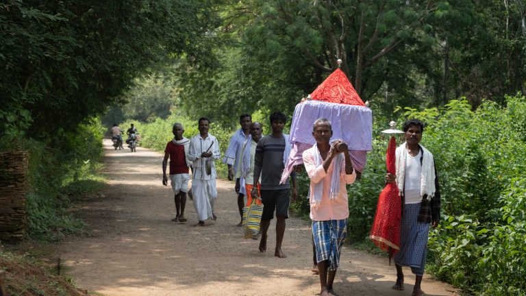 A photo of men carrying a coffin along a tree lined street.